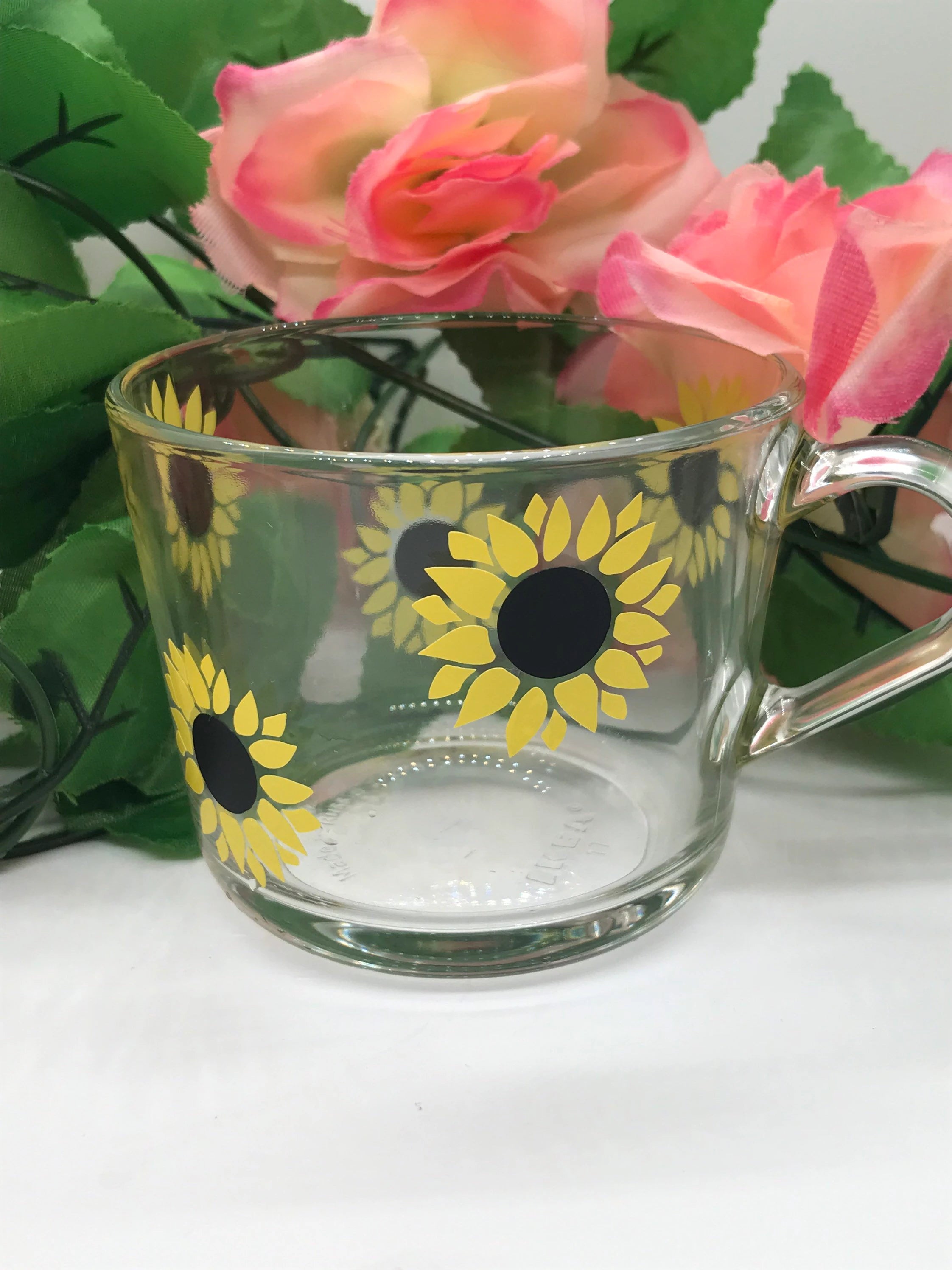 Clear glass mug with sunflower design on a white surface with pink flowers in the background