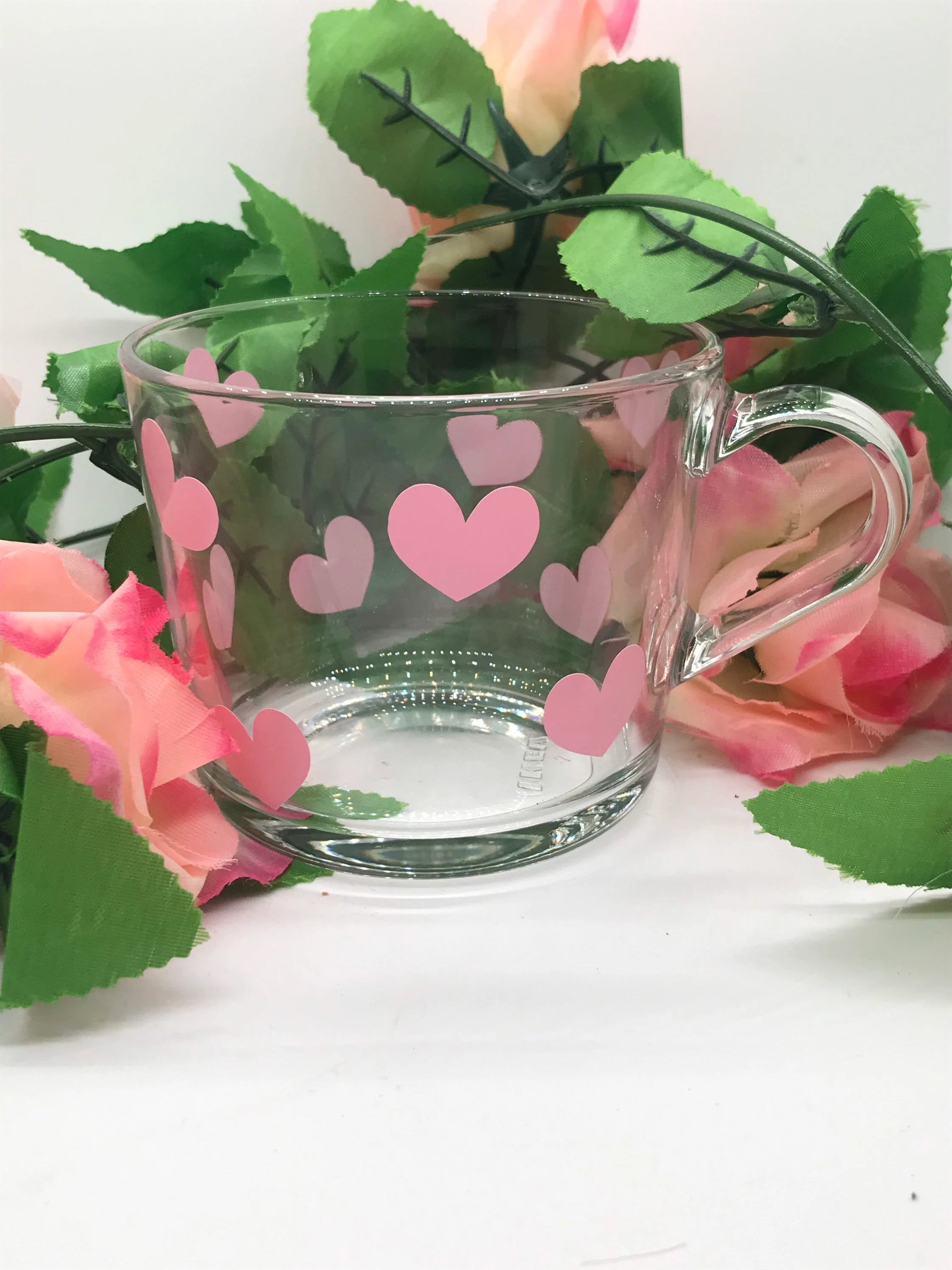 Clear glass mug with pink hearts on a white background with flowers and leaves.