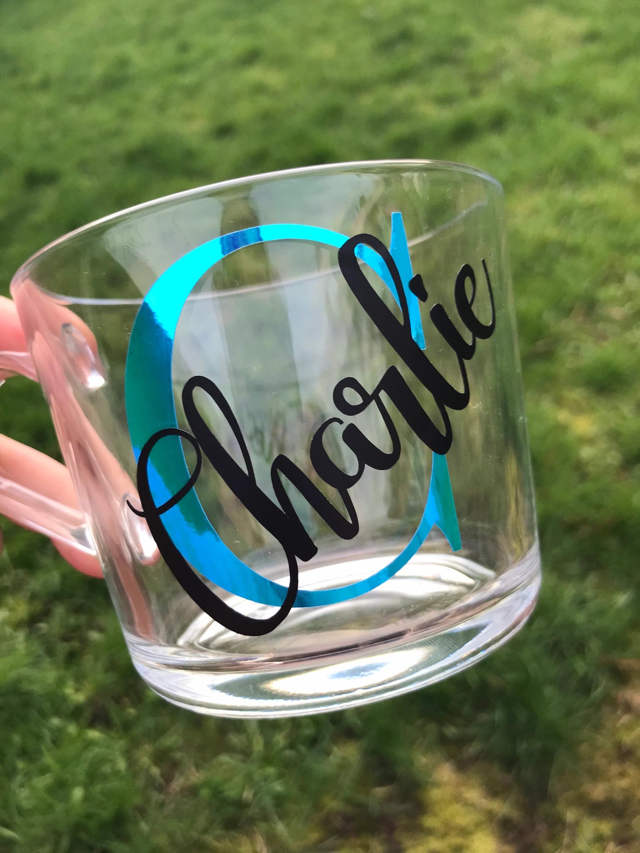 Clear glass mug with 'Charlie' printed on it, held by a hand against a grassy background