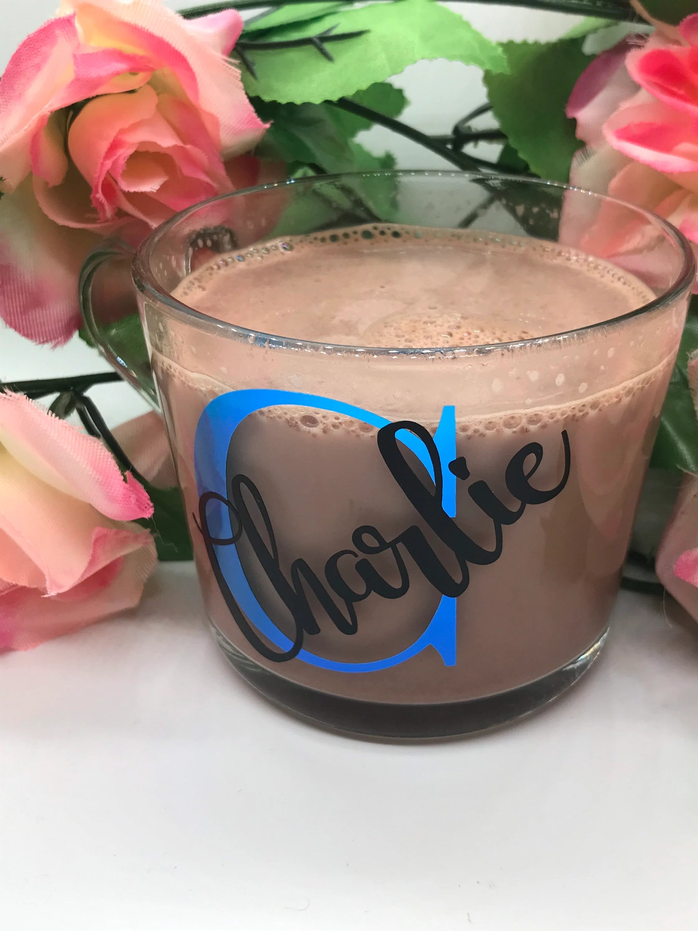 Glass mug with a name on it, filled with a hot chocolate, surrounded by pink flowers.