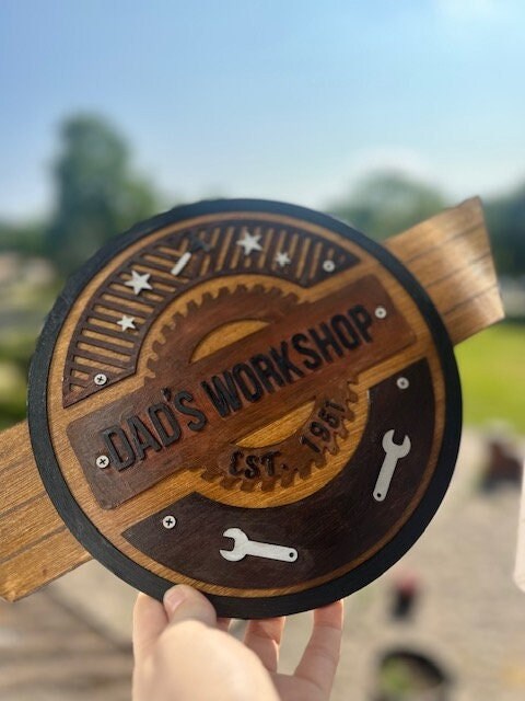 Wooden sign with 'Dad's Workshop' engraving held outdoors.