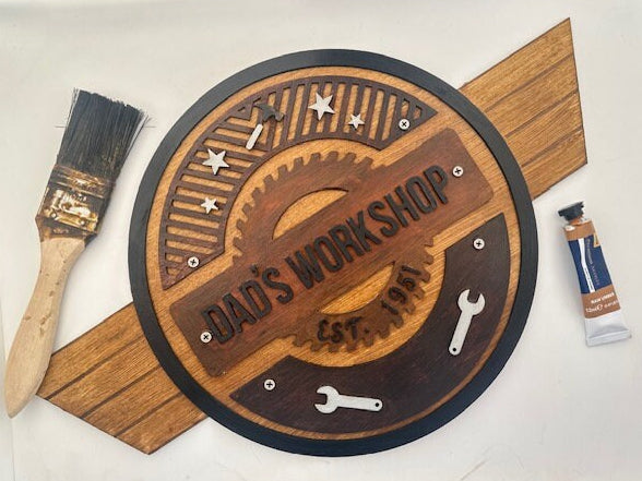 Wooden sign with 'Dad's Workshop' engraving, paintbrush, and paint tube on a white background