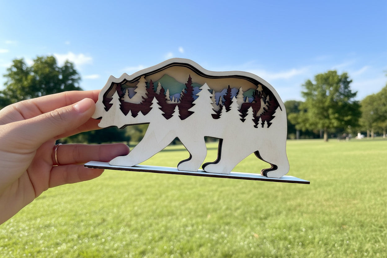 Hand holding a bear-shaped cutout with forest design against a grassy field background