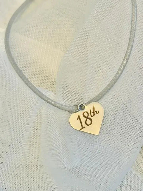 Heart-shaped pendant with '18th' on a grey cord against a light fabric background