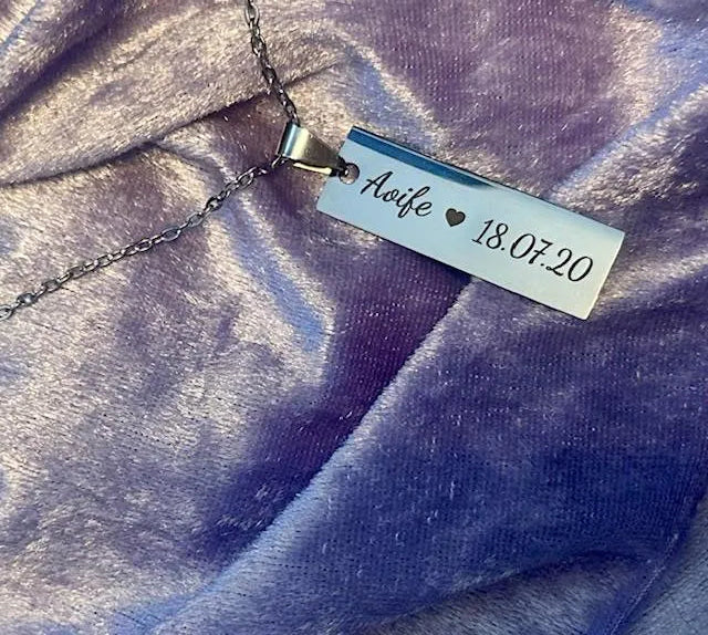 Necklace with a rectangular tag engraved with 'Aoife', a heart, and '18.07.20' on a purple fabric background.