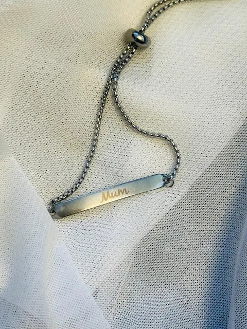 Silver stainless steel bracelet with a bar pendant engraved with 'Mum' on a textured fabric background