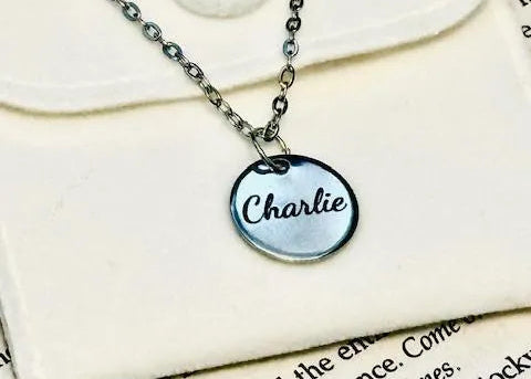 Stainless steel silver necklace with a round pendant engraved with 'Charlie' on a textured surface.